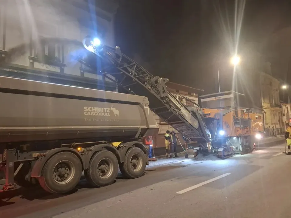 Un utilaj de construcții cu o bandă transportoare ridicate descarcă materiale într-un camion mare pe un drum asfaltat noaptea.