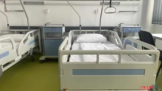Două paturi de spital cu lenjerie albă și mese de noapte albe cu sertare albastre sunt amplasate într-o cameră cu podea verde.