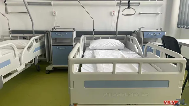 Două paturi de spital cu lenjerie albă și mese de noapte albe cu sertare albastre sunt amplasate într-o cameră cu podea verde.