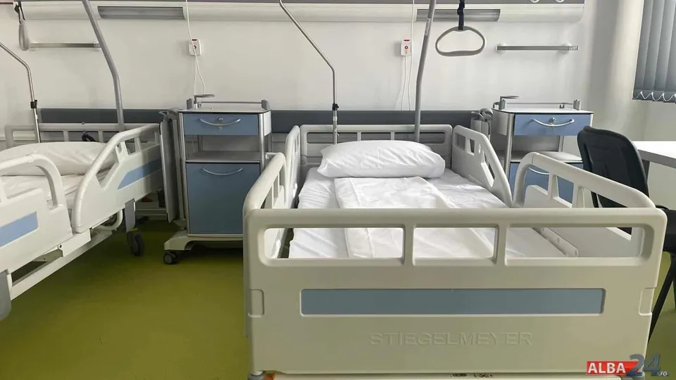 Două paturi de spital cu lenjerie albă și mese de noapte albe cu sertare albastre sunt amplasate într-o cameră cu podea verde.