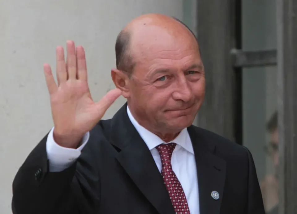 Un bărbat cu părul rare, purtând costum negru, cămașă albă și cravată roșie cu puncte albe, ridică mâna dreaptă în semn de salut.