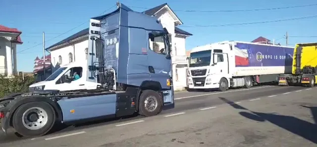 Mai multe camioane și TIR-uri sunt parcate pe o stradă asfaltată lângă clădiri sub un cer senin.