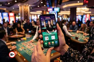 O persoană ține un smartphone care afișează un joc de blackjack live, în timp ce în fundal se vede un casino cu mese de joc.