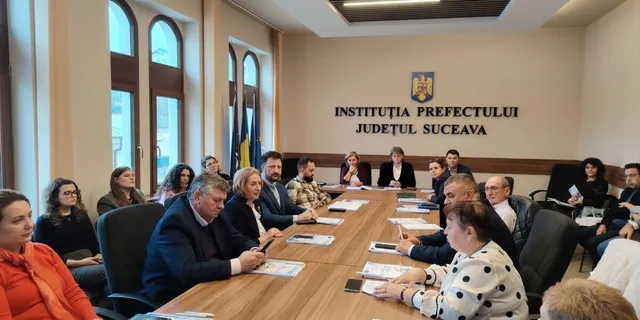 O grupă de persoane stă la o masă lungă de lemn într-o sală de ședințe, cu documente și telefoane mobile pe suprafață.