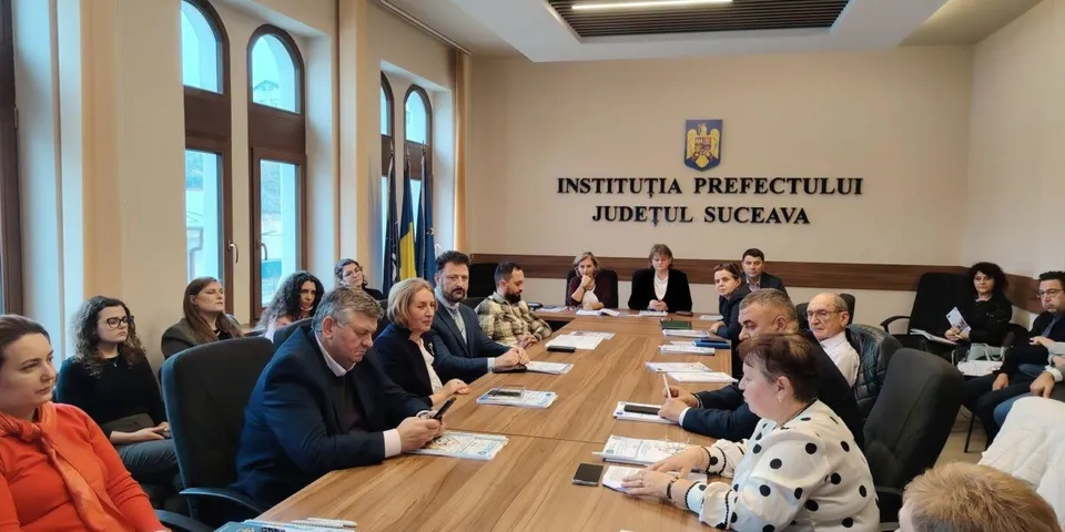 O grupă de persoane stă la o masă lungă de lemn într-o sală de ședințe, cu documente și telefoane mobile pe suprafață.