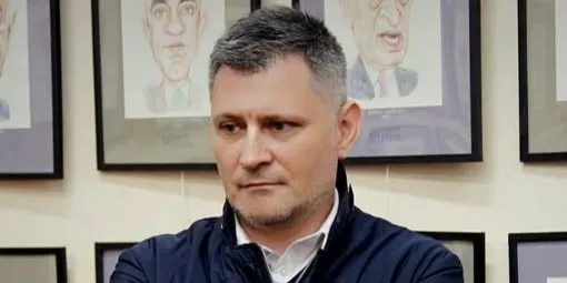 Un bărbat cu părul scurt și grijî, purtând o cămașă albă și o jachetă închisă la culoare, stă în fața unor tablouri înrămate pe un perete.