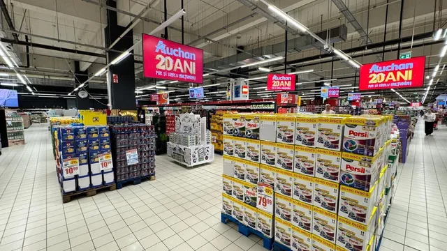 Rânduri de rafturi și stive de produse de curățenie în ambalaje de carton sunt dispuse pe podeaua unui magazin de alimente.