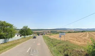 O stradă asfaltată cu linii albe marcate trece pe lângă un câmp brun și un semn de circulație alb cu text negru și bordură albastră.