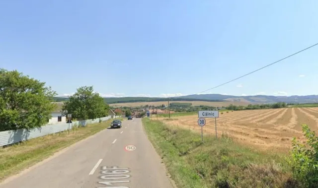 O stradă asfaltată cu linii albe marcate trece pe lângă un câmp brun și un semn de circulație alb cu text negru și bordură albastră.