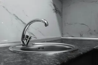 Un robinet metalic de bucătărie este montat pe o chiuvetă rotundă de inox, situată pe un blat de culoare închisă lângă un perete marmorat.