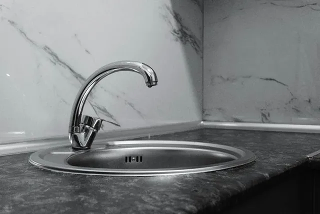 Un robinet metalic de bucătărie este montat pe o chiuvetă rotundă de inox, situată pe un blat de culoare închisă lângă un perete marmorat.