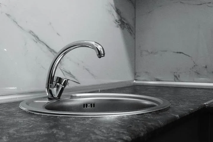 Un robinet metalic de bucătărie este montat pe o chiuvetă rotundă de inox, situată pe un blat de culoare închisă lângă un perete marmorat.