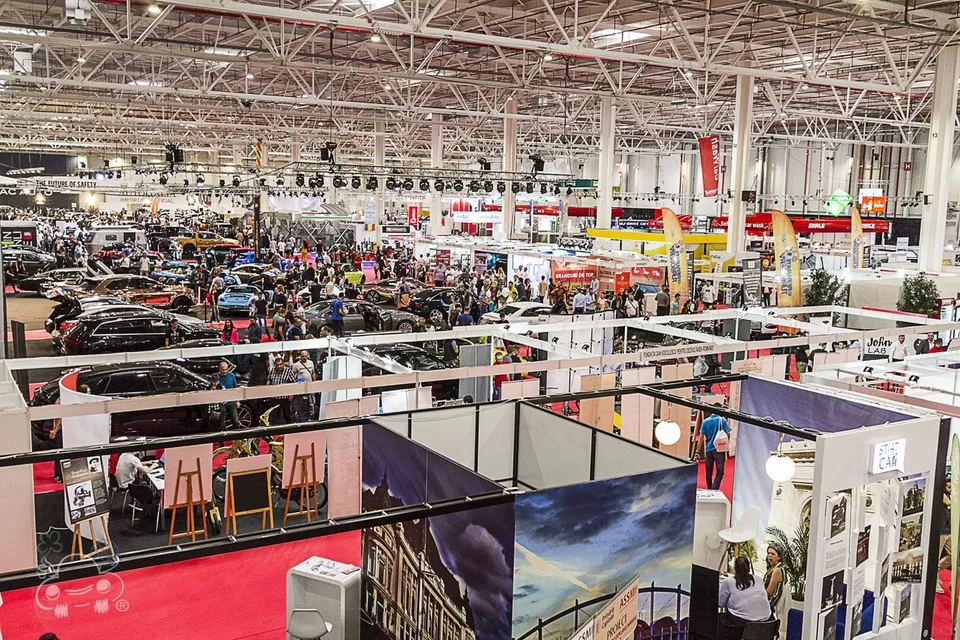 O perspectivă de sus asupra unui spațiu de expoziție mare cu standuri, mașini parcate și mulțimi de oameni circulând printre ele.