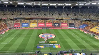 Un teren de fotbal verde cu linii albe este situat în interiorul unui stadion mare cu tribune pline de spectatori sub lumină artificială.