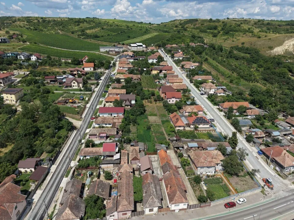 O vedere aeriană prezintă o zonă rezidențială cu case cu acoperișuri înclinate, drumuri și dealuri verzi sub un cer cu nori.