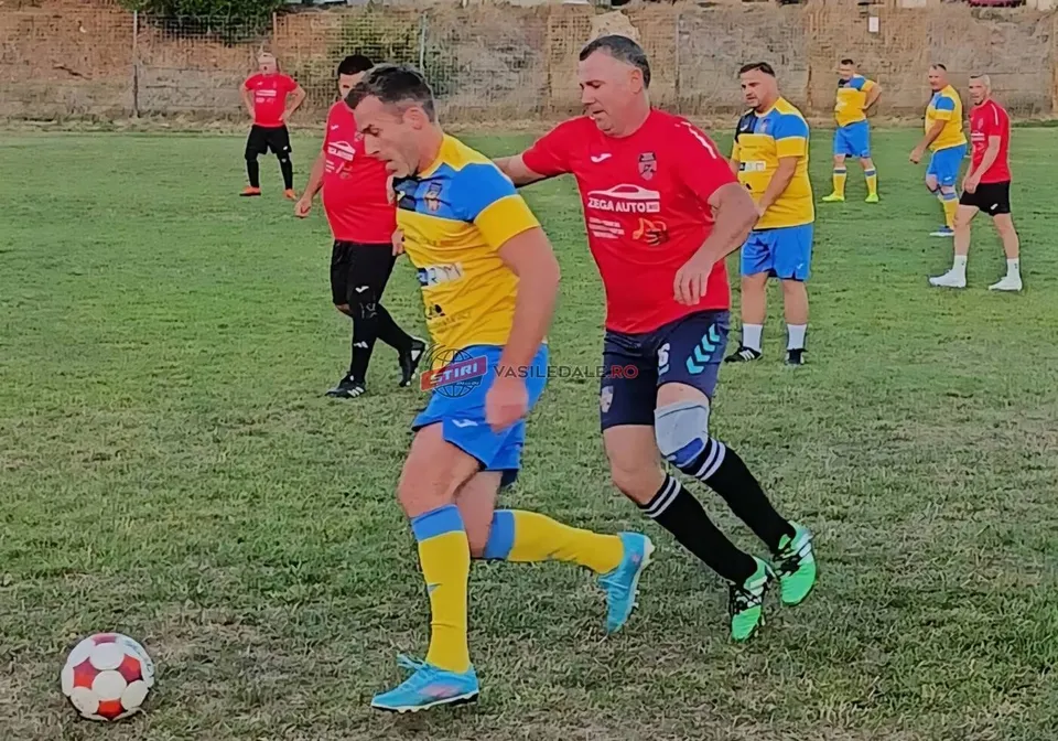 Un jucător în echipament galben și albastru conduce un fotbal pe un teren de iarbă, fiind urmărit de un adversar în echipament roșu.
