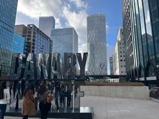 Mai mulți oameni stau pe o terasă lângă un semn metalic mare cu textul CANARY, în fața unor clădiri înalte de birouri.
