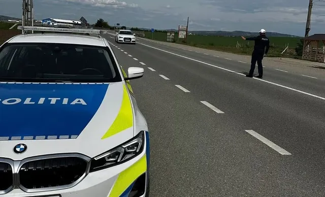 Un autoturism inscripționat Poliția este oprit lângă un drum, iar o persoană în uniformă stă la marginea carosabilului.
