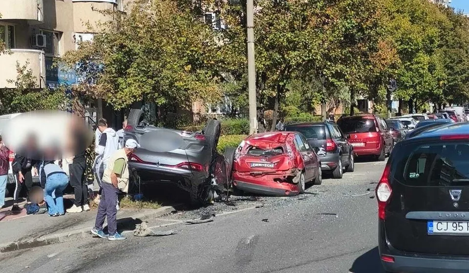 Un autoturism negru este răsturnat pe o parte pe marginea drumului, lângă un autoturism roșu avariat și alte vehicule parcate pe stradă.