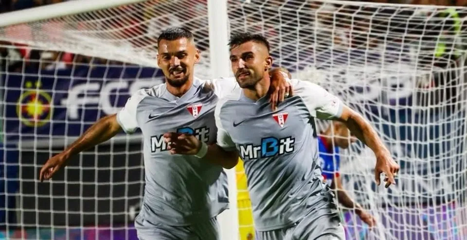 Doi jucători de fotbal în echipamente gri sărbătoresc în fața unei porți de fotbal sub luminile unui stadion.