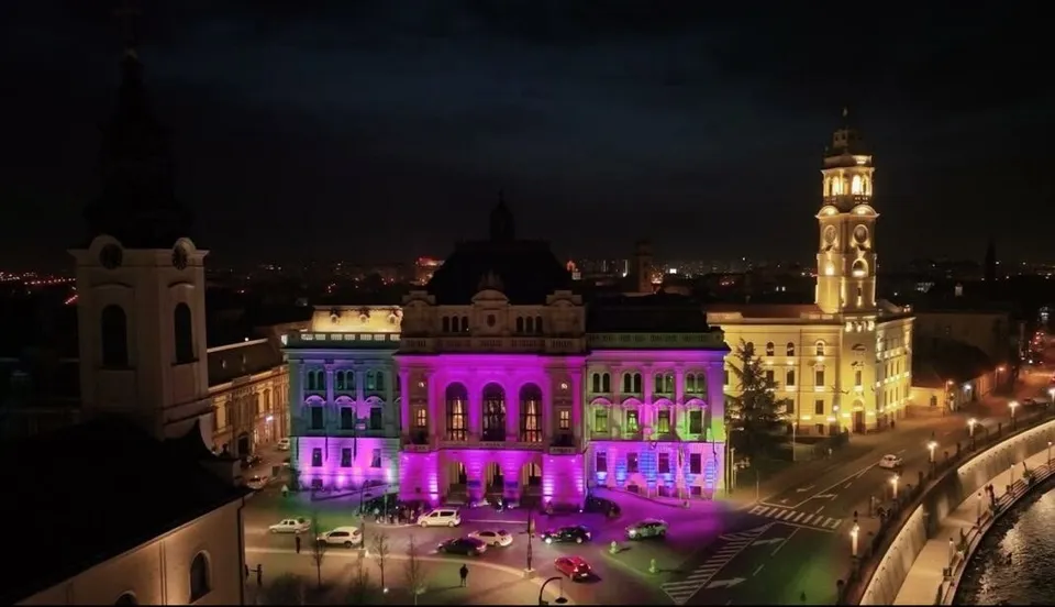 O clădire mare este iluminată cu lumini roz și mov pe fațadă, fiind situată lângă un turn înalt și o stradă cu mașini parcate.