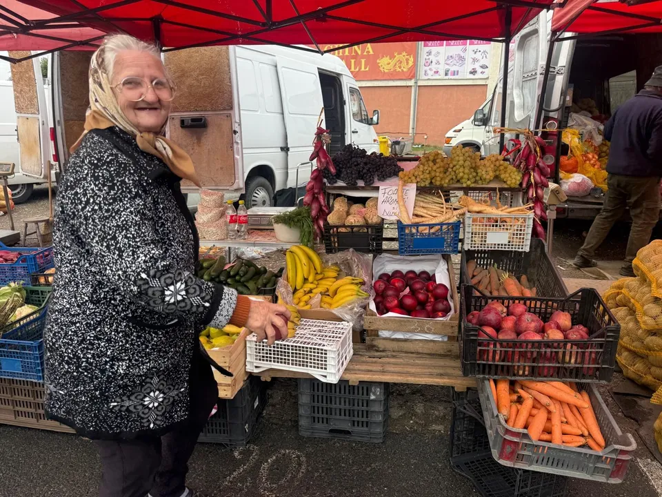 O femeie în vârstă, cu un eșarfa pe cap și o jachetă cu model, zâmbește într-o piață cu fructe și legume expuse pe mese, sub un acoperiș roșu.