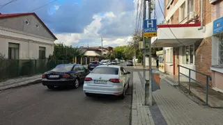 Două mașini sunt parcate pe marginea unui drum asfaltat lângă un trotuar și o clădire cu un panou albastru marcat cu litera H.