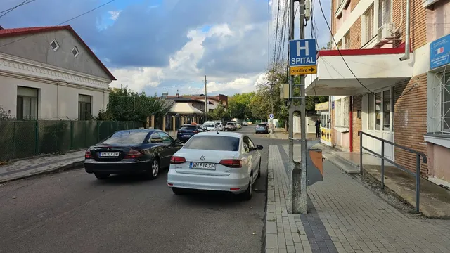 Două mașini sunt parcate pe marginea unui drum asfaltat lângă un trotuar și o clădire cu un panou albastru marcat cu litera H.