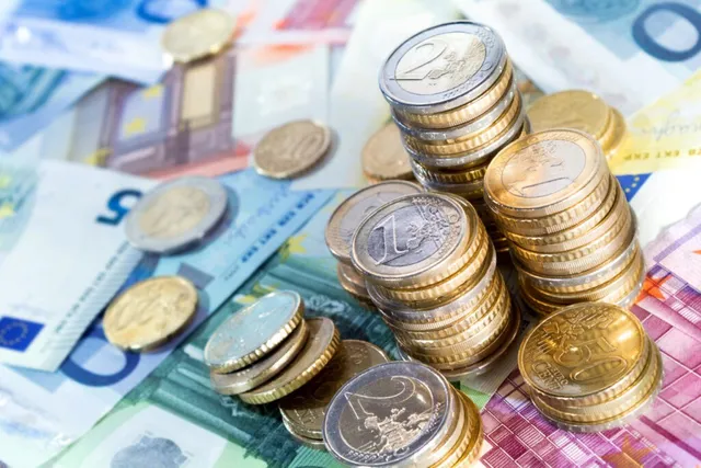 Mai multe stive de monede euro și monede individuale sunt împrăștiate pe diverse bancnote colorate.