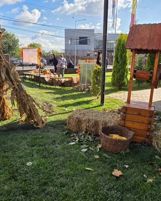 Oamenii stau lângă mese cu produse de toamnă, un puț de lemn, un coș cu fructe și un cub de fân pe iarbă.