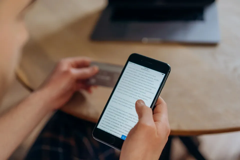 O persoană ține un smartphone cu un text afișat pe ecran și un card de plăți în mâinile sale deasupra unei mese de lemn.