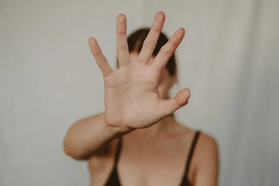 O persoană cu părul închis își întinde palma în față, acoperind parțial fața, în fața unui fundal neutru și deschis la culoare.