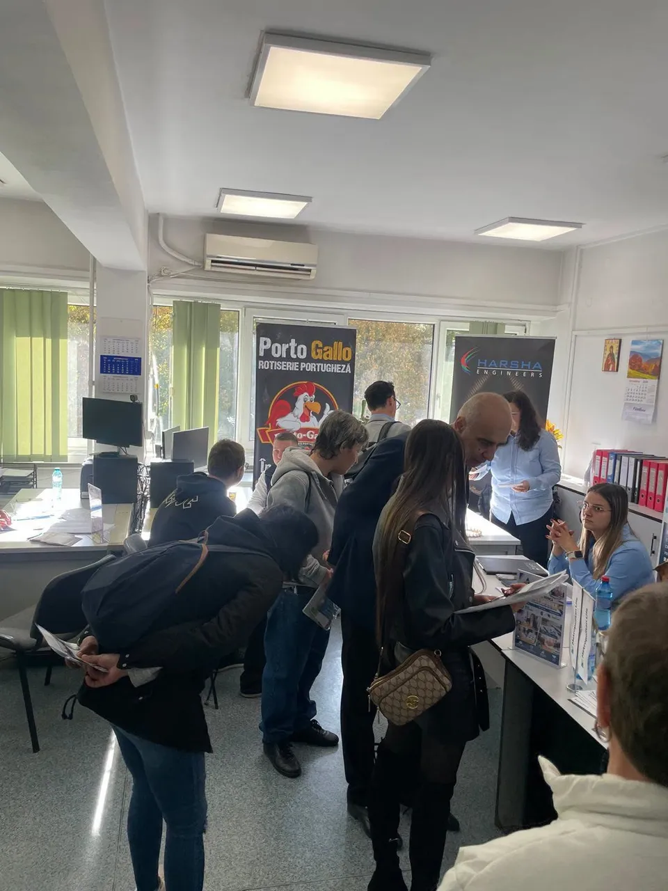 Oamenii stau în picioare într-o încăpere cu mese, consultând documente lângă standuri de prezentare cu afișe și monitoare de calculator.