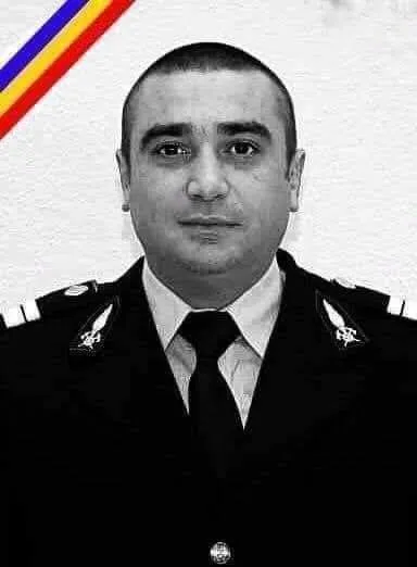 Un bărbat cu părul scurt poartă o uniformă închisă la culoare, cămașă deschisă la culoare și cravată, având insignele pe epoleți.