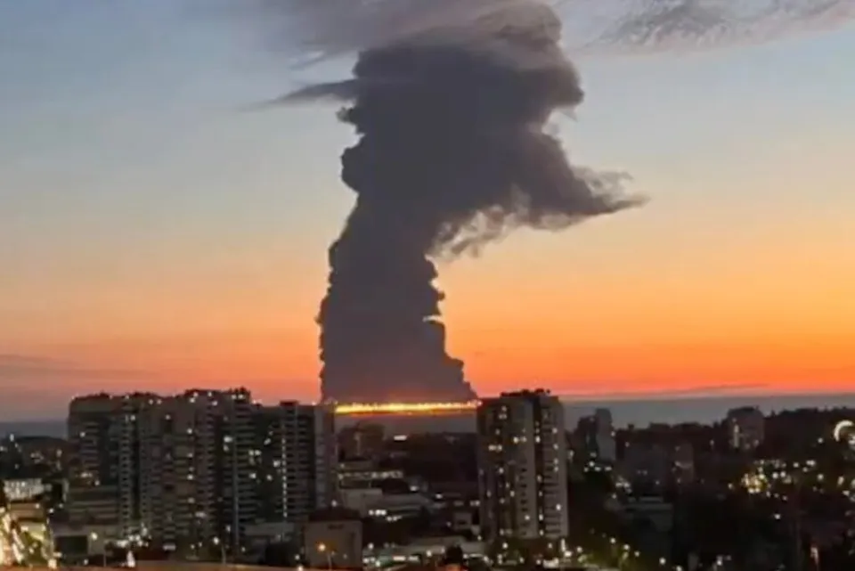 O coloană densă de fum negru se ridică deasupra unei linii de foc la orizontul mării, în fața unor clădiri urbane sub un cer la apus.