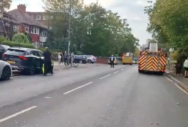 O stradă cu mașini parcate, un camion de pompieri cu marcaje galben-roșu și câteva persoane pe trotuar lângă un cordon de poliție.