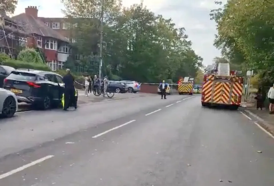 O stradă cu mașini parcate, un camion de pompieri cu marcaje galben-roșu și câteva persoane pe trotuar lângă un cordon de poliție.