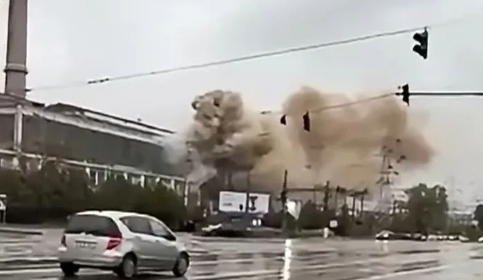 Un nor dens de fum și praf se ridică deasupra unor structuri industriale lângă o stradă umedă cu un autoturism alb în prim-plan.