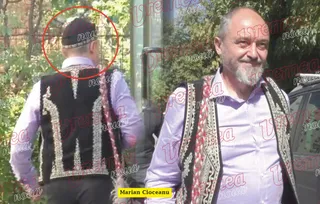 Un bărbat cu barbă și vestă tradițională neagră stă în picioare lângă un alt bărbat care poartă vestă tradițională și este văzut din spate.