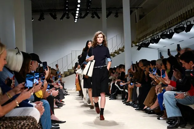 O femeie cu părul lung și ondulat merge pe o pistă de catwalk purtând un tricou închis la culoare, pantaloni scurți negri și o geantă albă.
