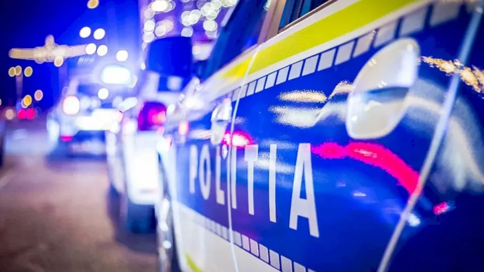 Un vehicul de poliție albastru și alb cu inscripția POLIȚIA este surprins în prim-plan, fiind luminat de lumini strălucitoare pe un fundal nocturn.