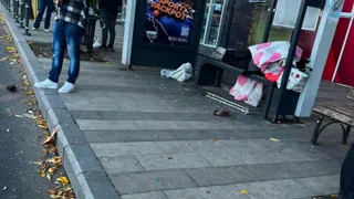 O persoană stă în picioare lângă o stație de autobuz unde sunt împrăștiate obiecte și pungi pe podeaua pavată.