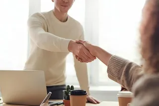 Un bărbat într-un pulover deschis se strânge în mână cu o persoană aflată în prim-plan, lângă un laptop și două pahare de cafea pe masă.