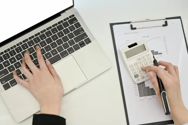 Mâinile unei persoane tastând pe un laptop și folosind un calculator lângă un dosar cu grafice pe o masă albă.