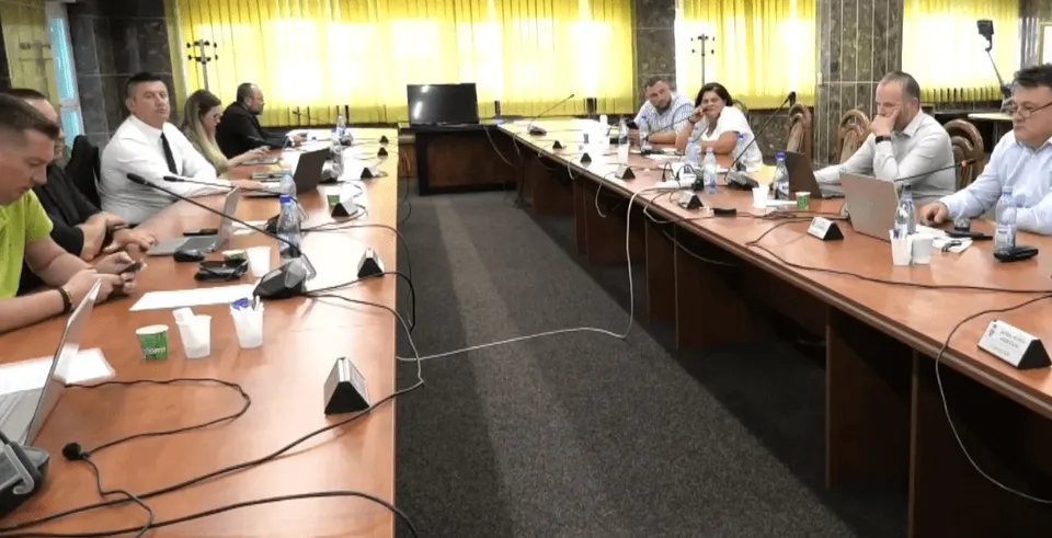 Oamenii stau la mese lungi de conferință într-o încăpere cu ferestre mari, având în față laptopuri, telefoane și microfoane pe suprafața de lemn.