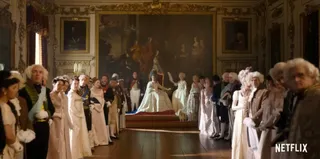 Un grup de persoane îmbrăcate în haine de epocă stau în picioare într-o sală decorată cu tablouri și detalii aurii, în jurul unei femei așezate pe un fotoliu.