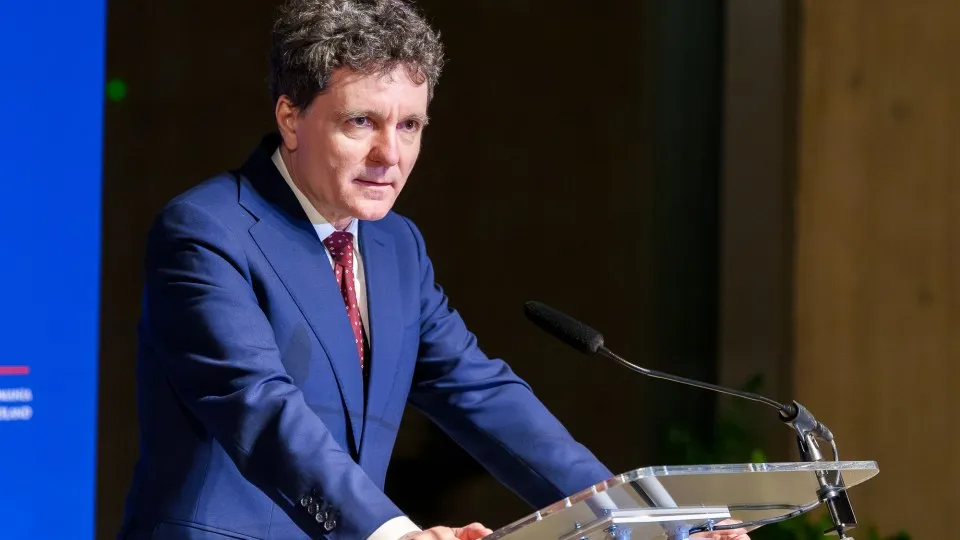 Un bărbat cu părul creț, îmbrăcat în costum albastru și cravată roșie, stă în spatele unui pupitru transparent lângă un microfon.
