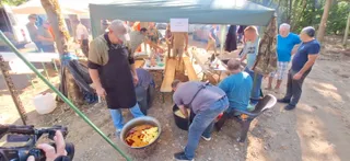 Un grup de bărbați stă sub un prelat verde lângă mese cu mâncare și un vas mare cu preparat culinar.