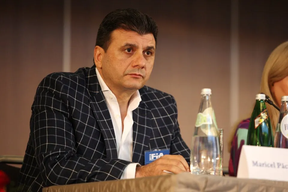 Un bărbat cu părul scurt și sacou în model carou, purtând o cămașă albă, stă la o masă lângă sticle de apă.