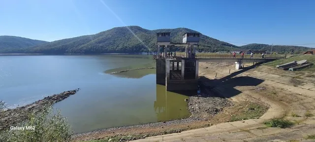 O structură de beton cu două turnuri albe se află lângă un lac, sub un cer senin și dealuri verzi.